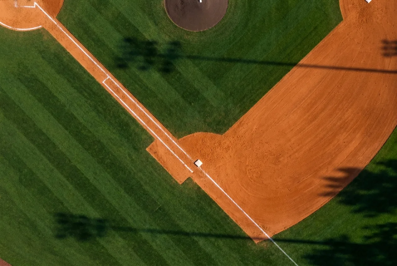 Aerial view of a groomed college-baseball infield corner — orange clay, green turf, a chalk foul line and a base.
